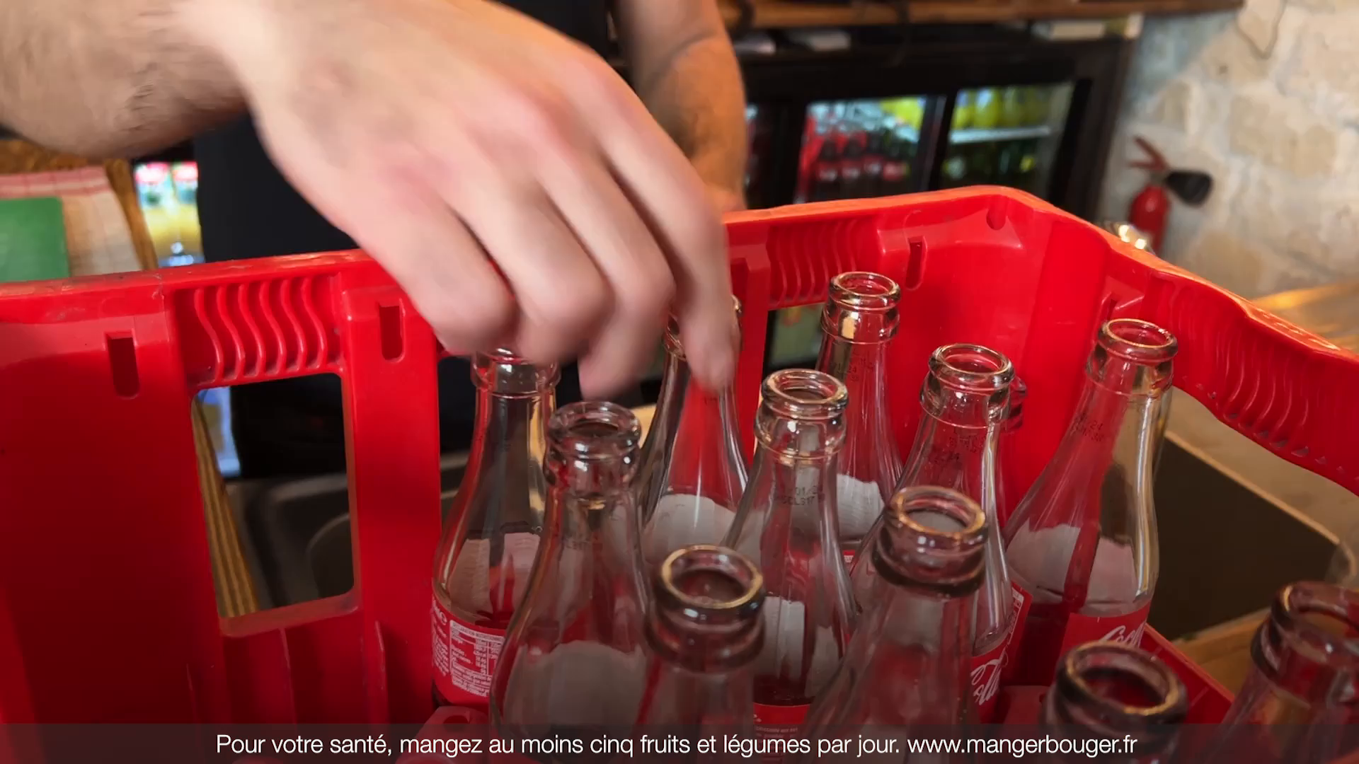 Main plaçant des bouteilles en verre consignées Coca‑Cola dans une caisse rouge dans un café, hôtel ou restaurant.