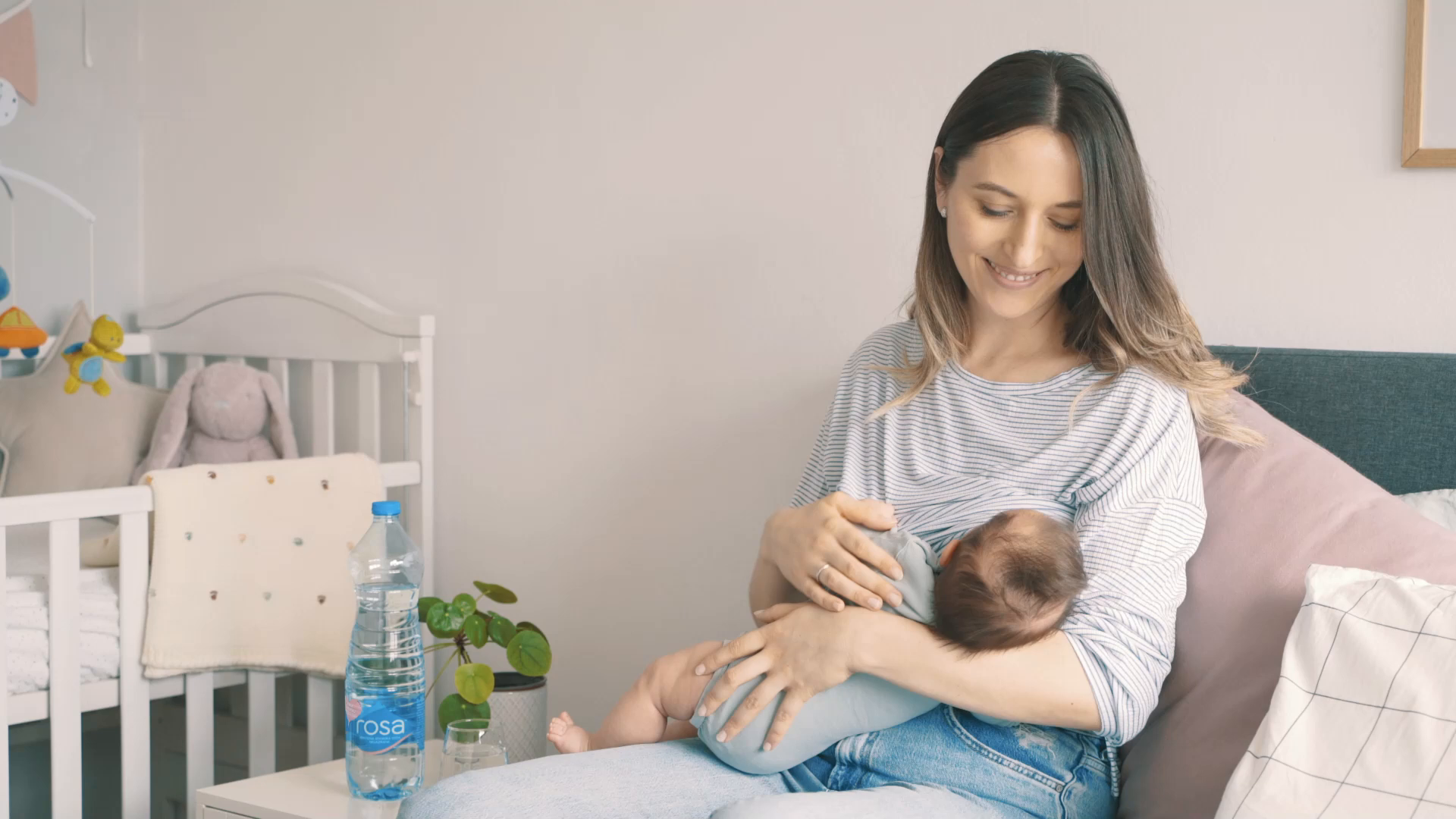 Žena sedi na krevetu i doji bebu, dok je pored nje boca i čaša Rosa vode na noćnom ormariću blizu dečijeg krevetića.